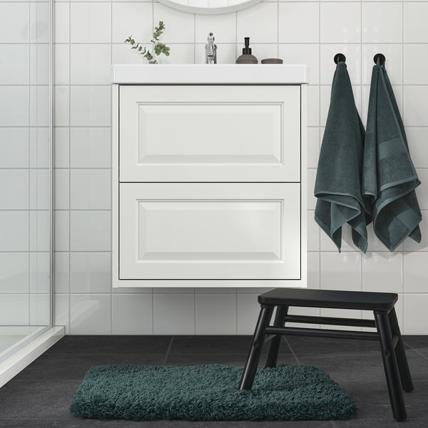 Minimalist bathroom featuring ALMTJÄRN teal bath mat, hanging towels, white cabinet, and black stool.