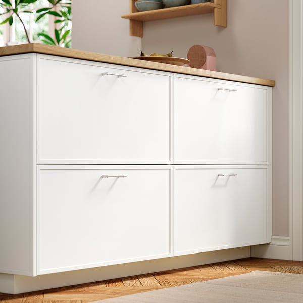 White kitchen cabinet with thin frame, four doors and a wooden countertop, mounted on a pale pink wall, with decorative items on top.