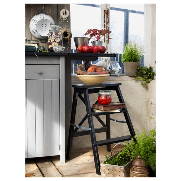 A black BEKVÄM step ladder near a wooden cabinet, holding a bowl of fruits and a jar.