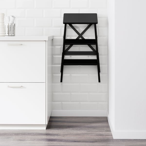 Black stepladder against white tiled wall in kitchen, folded for compact storage.