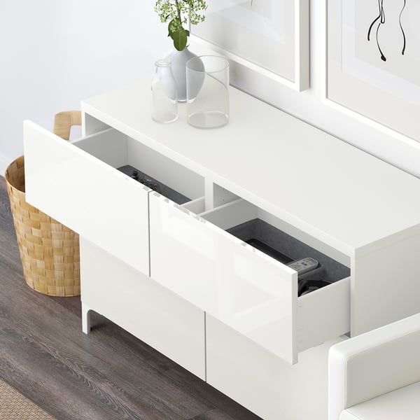 White BESTÅ storage unit with open drawers revealing remote controls. Decorative vases on top.