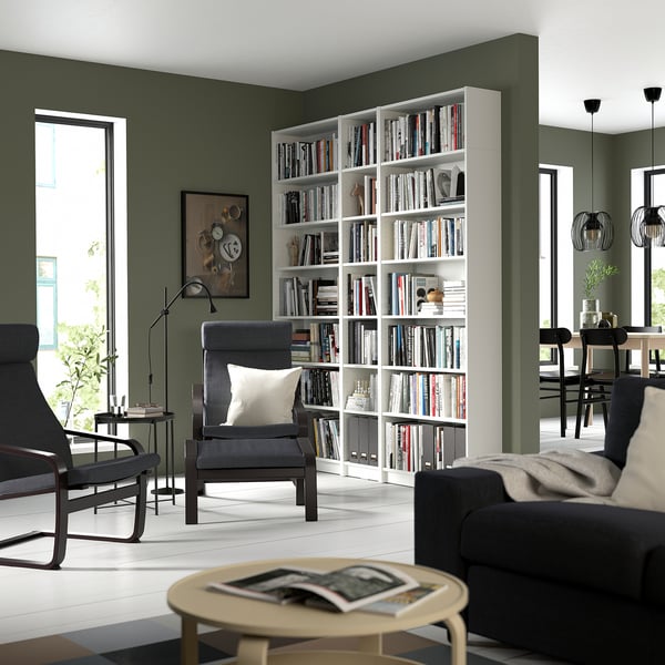 Modular white bookshelf filled with books, between green walls, with black furniture, creating a cosy reading nook.