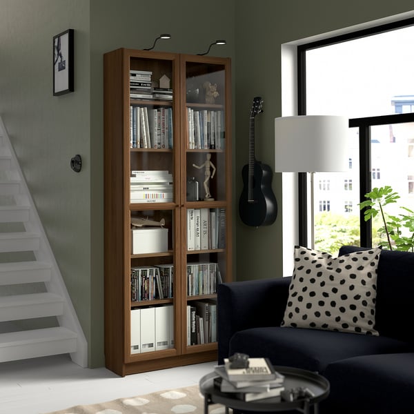 Modern living space with BILLY bookcase, navy sofa, and guitar. Shelves hold books and decor. White stairs and large window.