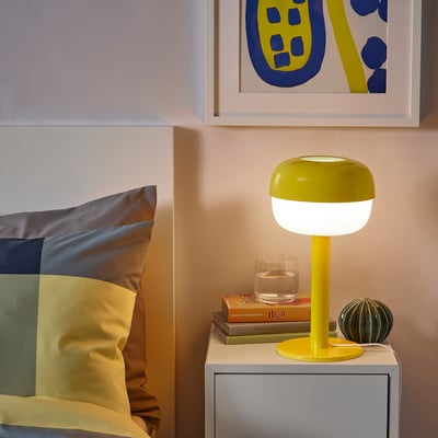 Bright yellow BLÅSVERK lamp on bedside table, small glass & books. Modern retro design.