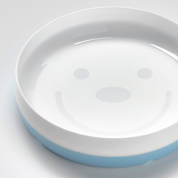 Round, white plate with blue edge, designed for kids. Smiling face, non-slip base, and angled sides for easy eating.