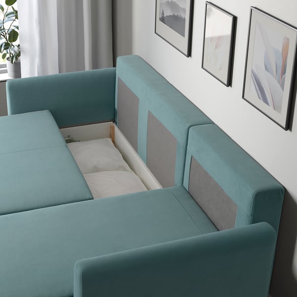 Teal sofa bed with storage, partly converted. Two pillows, grey art, curtain, plant.
