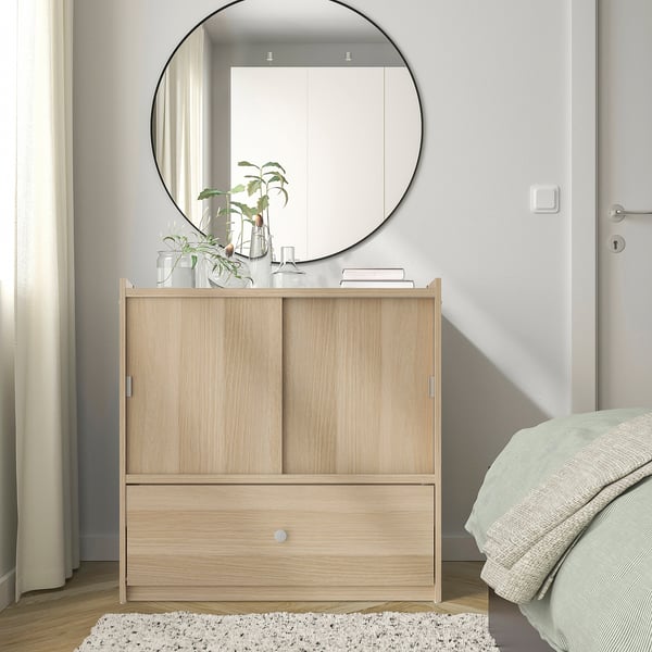 BRUKSVARA storage unit with light wood & sliding doors, smaller than bed & mirror, on rug near white bedspread.