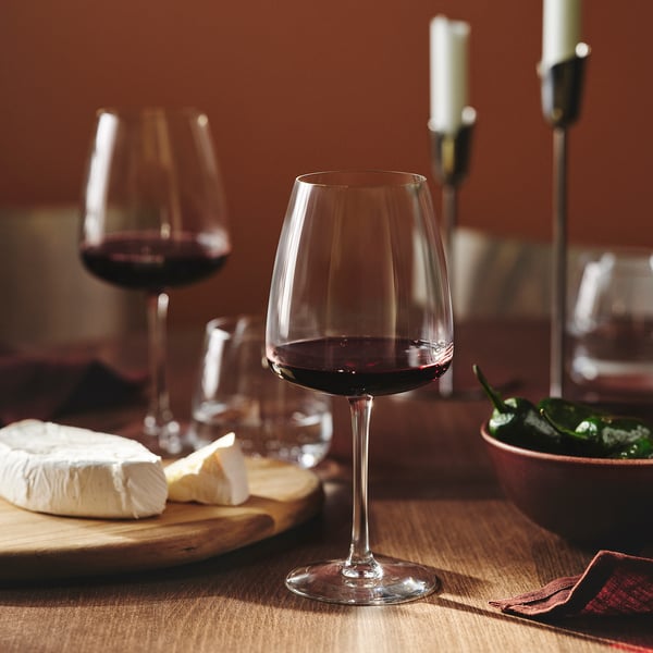 Clear wine glass with red wine stands on wooden table, surrounded by cheese and greens.
