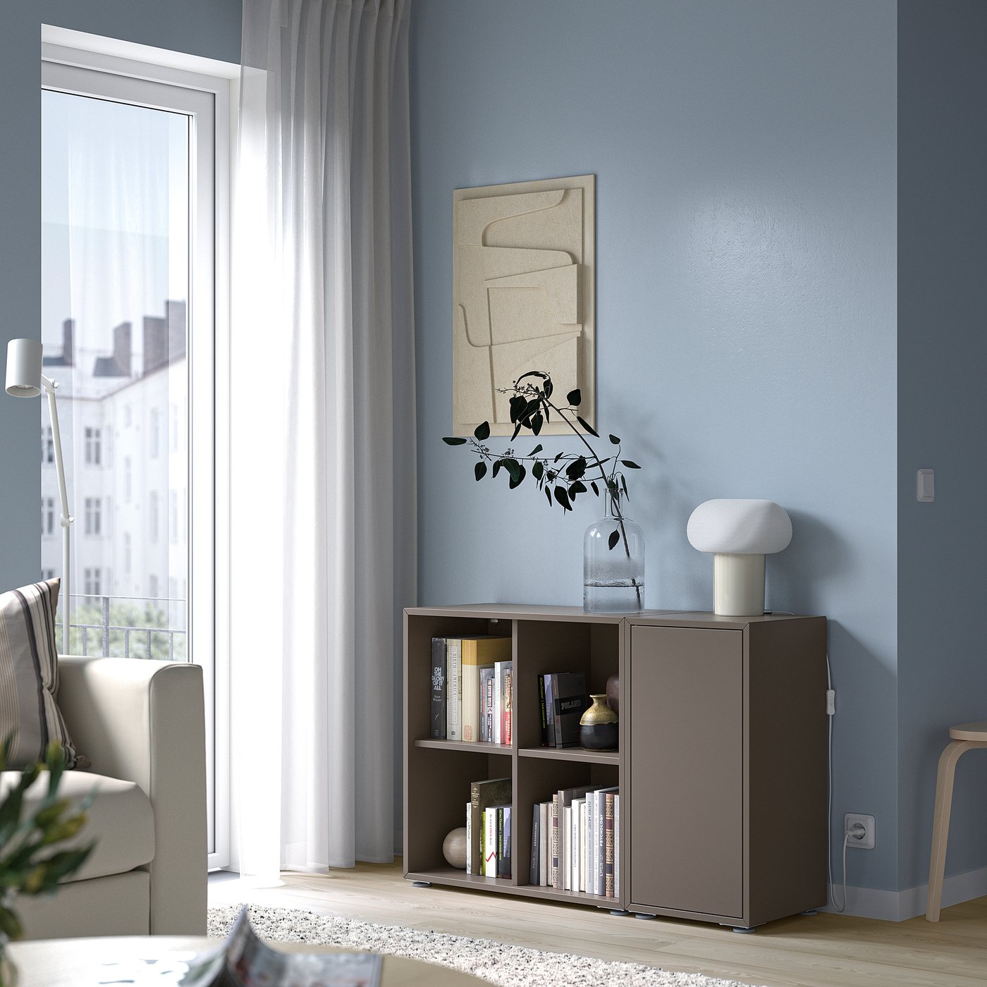 Modern living room with blue wall, abstract art, bookshelf, chair, and window with sheer curtains. Focus on EKET storage unit with books and vase.