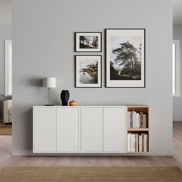 Modern living room with grey wall and wooden floor. White EKET cabinets, open shelves with books, table lamp, and wall art with nature photos.