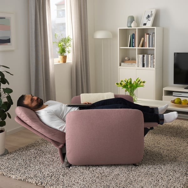 Person relaxes on adjustable pink EKOLSUND recliner in living room. Chair reclines, offering nap comfort. Nearby bookshelf, plants, flowers, and tv.