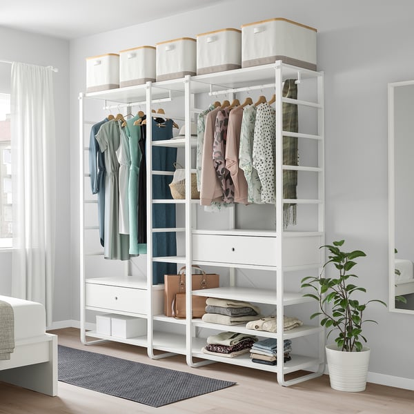 White ELVARLI storage system with shelves, drawers, and clothes. A plant adds nature. Tall next to bed.
