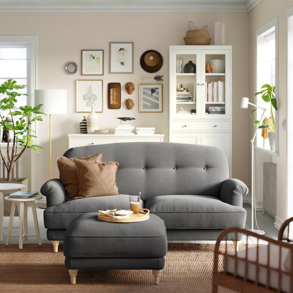 Modern living room with grey ESSEBODA sofa, brown pillows, wooden cabinet, plants, and pictures.