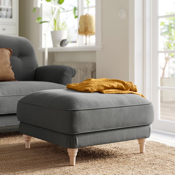 Grey footstool with wood legs, near blue sofa. Yellow blanket on top. Elegant, turned legs. Natural light streams in.