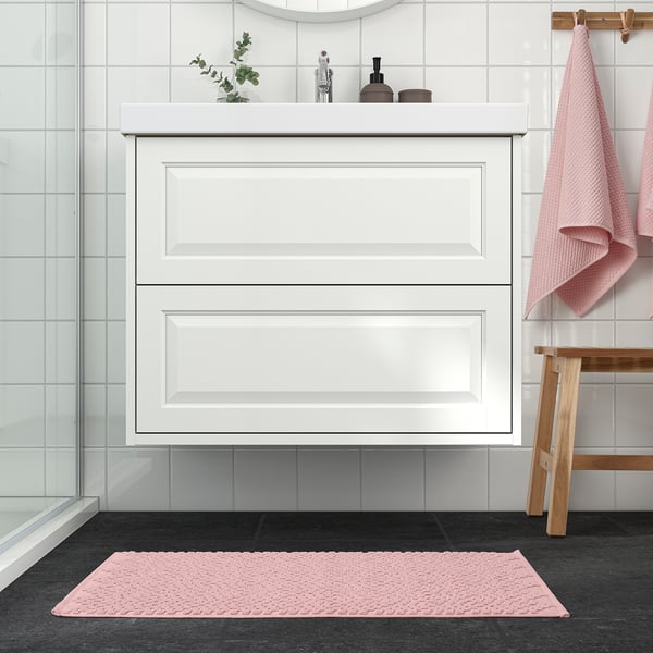 Modern bathroom with a pale pink FJÄLLKATTFOT bath mat. Mat fits neatly in front of white cabinet, complementing hanging towels and small plant.