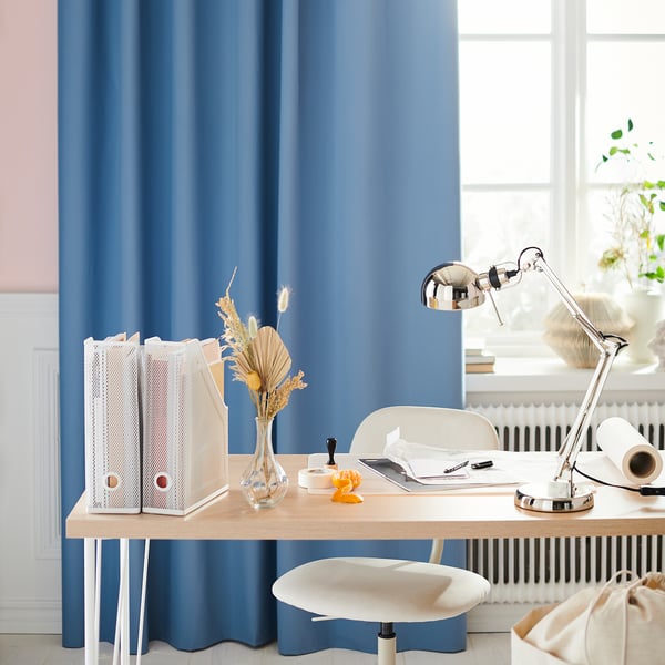 Modern home office with blue curtains, wooden desk, white chair, and adjustable silver FORSÅ lamp.