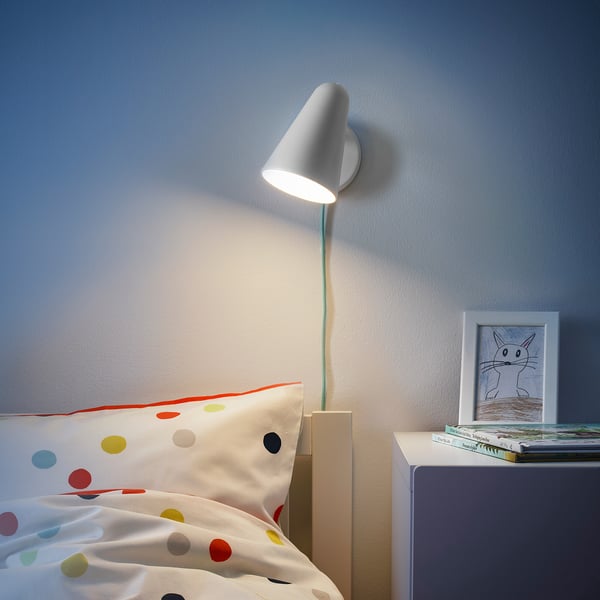 Modern wall-mounted lamp, white, conical shade, near colorful polka-dot bedding, nightstand with framed cat drawing, bright light.