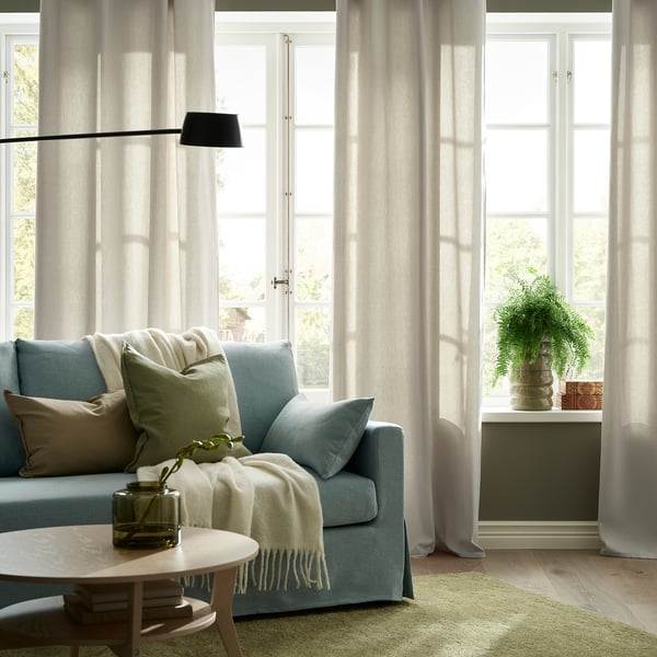 Cosy living room with large light beige GINSTMOTT curtains framing wide windows, complementing the blue sofa and adding a serene, private feel.