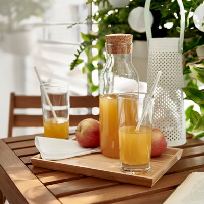 Two glasses of orange juice on a wooden tray with apples, napkins, and straws. Background has a lantern and green plants.