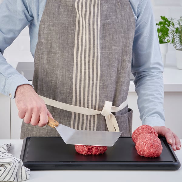 Person using a wide spatula to flip burgers on a tray. They wear an apron and striped shirt. The spatula is wooden-handled, ideal for grilling.