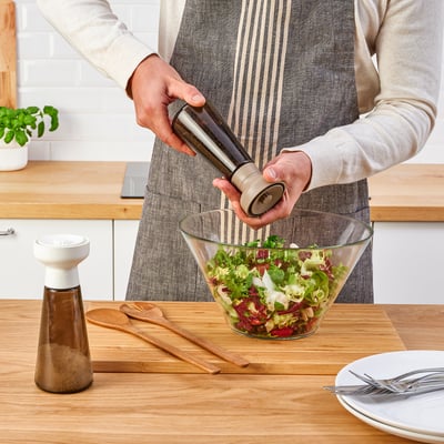 A person grinds pepper into a salad bowl with a HALVTOM spice mill on a wooden table.