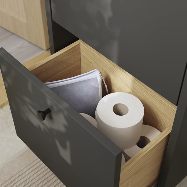 Bathroom cabinet with open drawer displaying toilet paper and other items. Dark grey finish with wood interior.