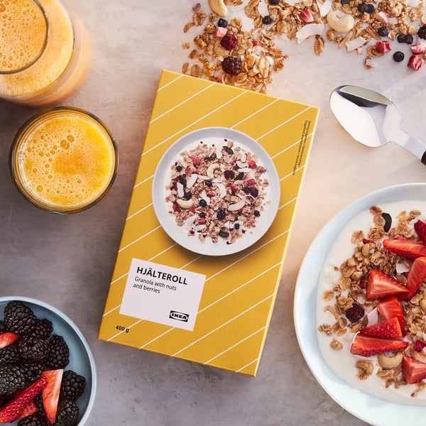 A bowl of HJÄLTEROLL granola featuring nuts and berries on a table with orange juice and yoghurt. The package is open, showing the granola inside.