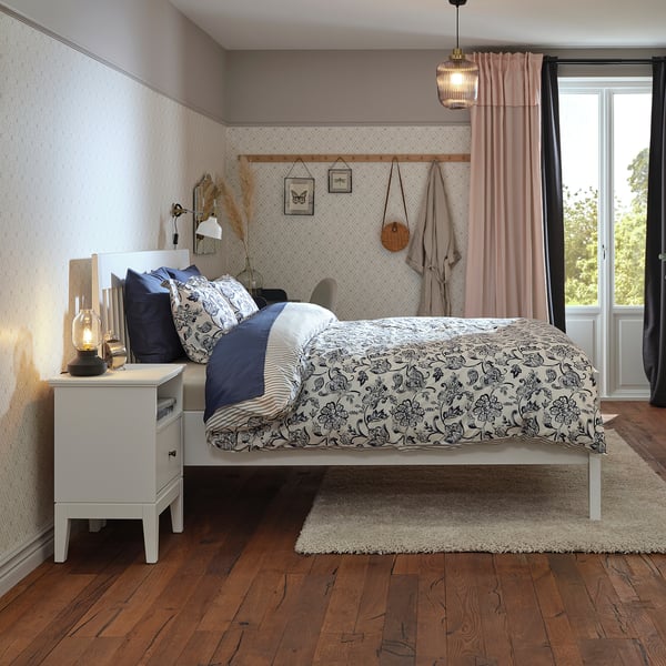 Cosy room with white IDANÄS bed, black & white floral bedding, wood floors, pink curtains, hanging rattan chair, white bedside table, lamp.