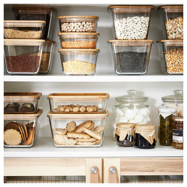 Pantry shelf with clear, square glass jars containing various dry foods. Lids are wooden. Jars are neatly arranged on white shelves.