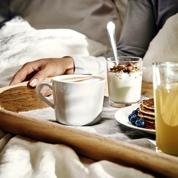 Hand holds mug of frothy coffee on tray with yoghurt, juice, and pancakes.