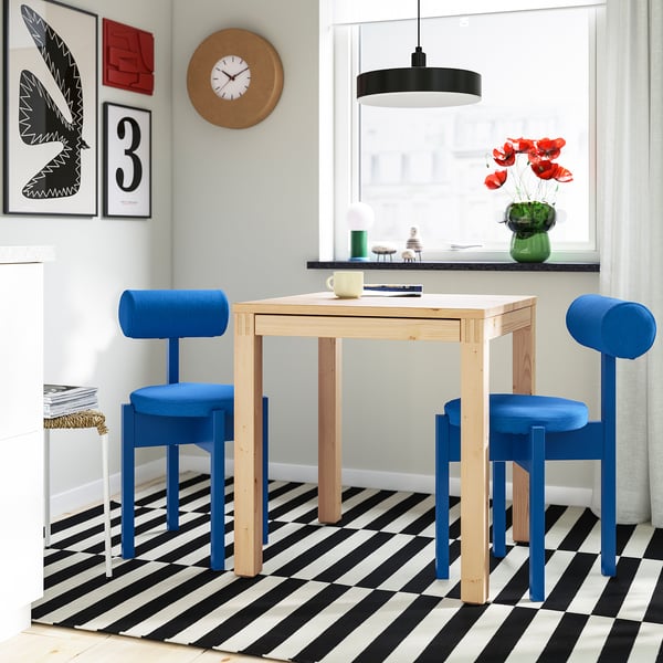 IKEA PS 2026: bright blue, cushioned chairs surround a light wood table. Black-and-white chequered floor. Bright, modern room setting.