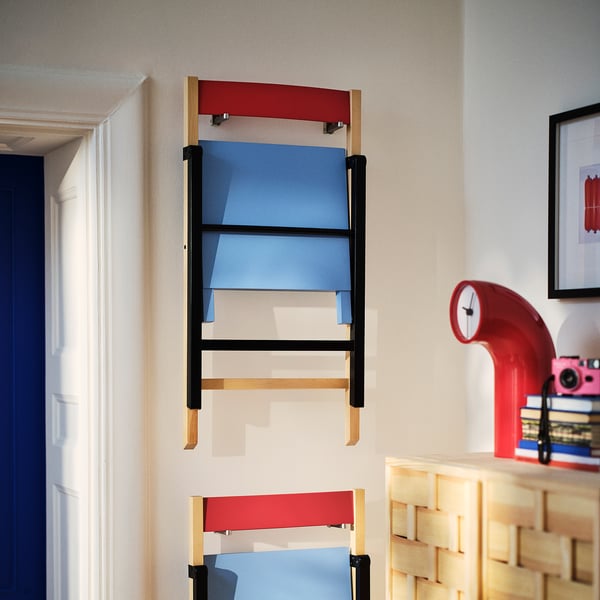 Modern wall-mounted foldable chair in bright red and blue with black metal supports and wooden legs. Suitable for compact spaces.