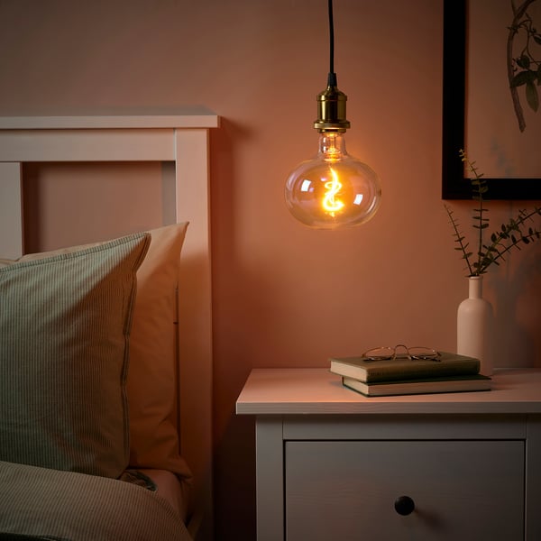 Bedroom with warm JÄLLBY lamp over bedside table with book and vase, bed with two pillows, peach walls.