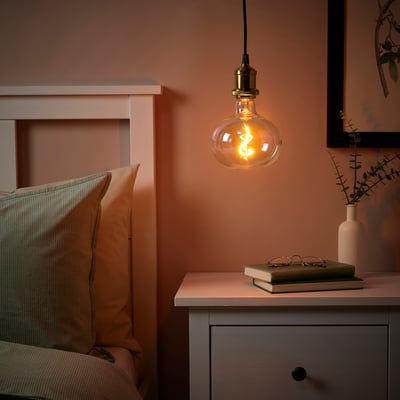 Bedroom with warm JÄLLBY lamp over bedside table with book and vase, bed with two pillows, peach walls.