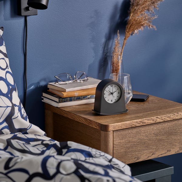 Bedside table with clock, glasses, books, phone, and vase. Clock shows time with luminous tips, rocks to wake up.