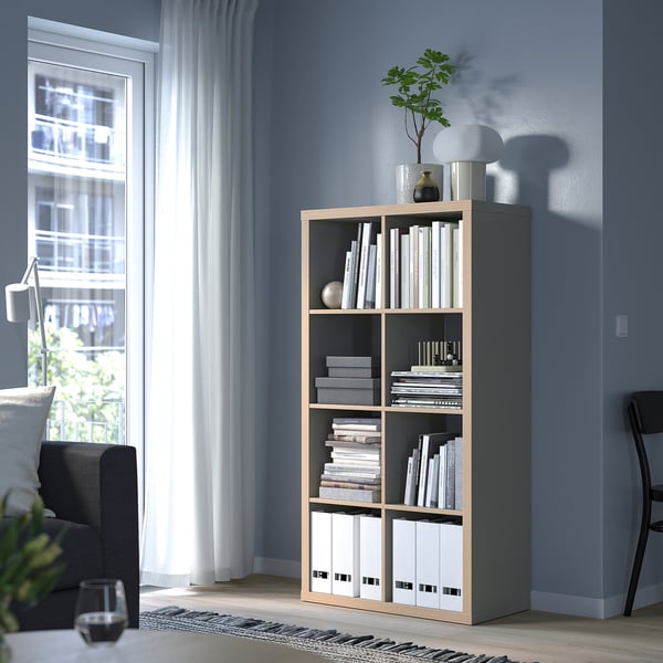 KALLAX bookshelf with books and decor, beside grey couch and black chair, against blue wall.