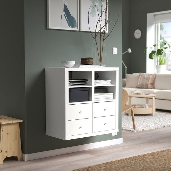 A white KALLAX unit against a green wall with shelves and drawers, displaying various items. A cosy living room setting, noticeable clock and plants.