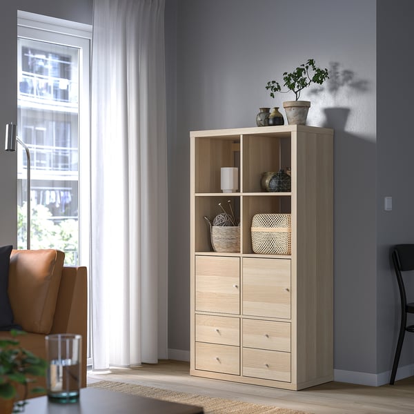 Wooden KALLAX shelf with drawers, vases, and plants beside window. Contrasts dark wall, central in bright room.