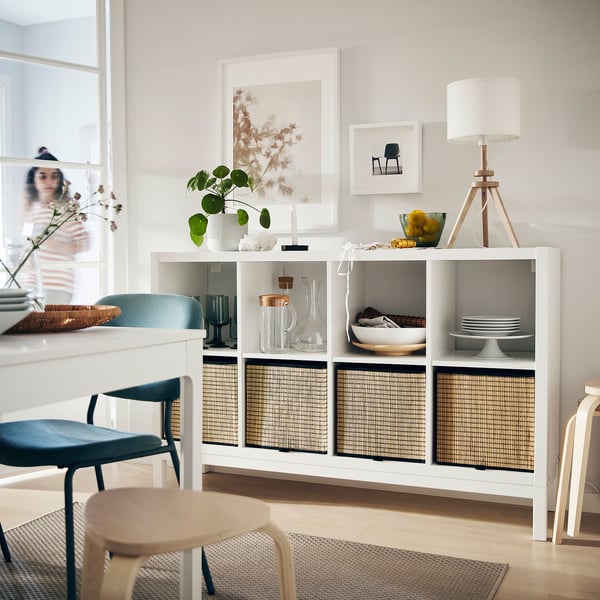 White KALLAX shelving unit with woven baskets, decorative items, and a table lamp, set against a wall with framed art.