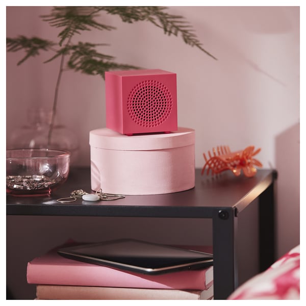 A stylish pink portable speaker sits on a white box atop a black shelf. Surrounded by decor items, it adds a vibrant touch to the setting.