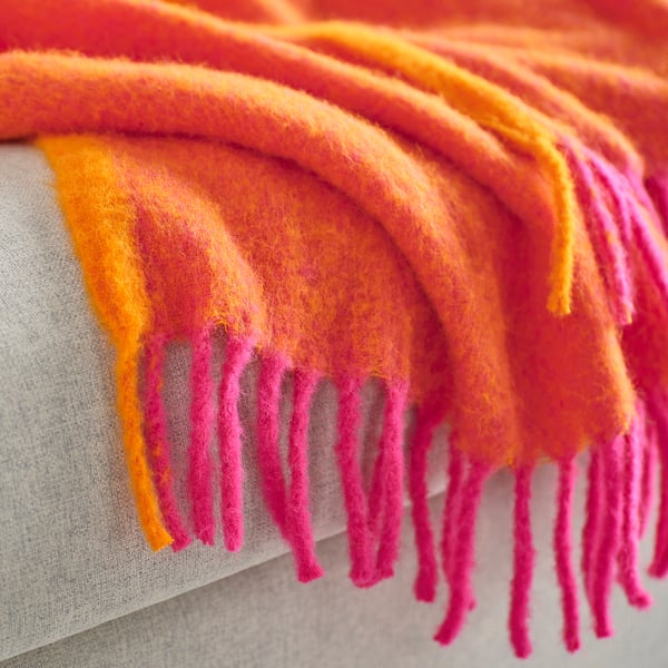 Bright pink and orange throw blanket with soft, fluffy texture, draped over edge of furniture, showing fringed ends.
