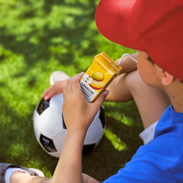 Child drinks yellow lang lur smoothie, with football ball, wearing red cap.