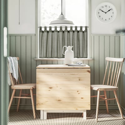 Wooden table with 2 chairs, green walls, grey curtains, window, white clock, pitcher.