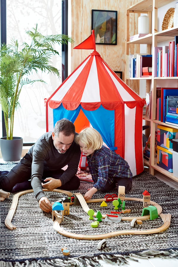 A person and child play with wooden LILLABO train set, red and white tent, bookshelves and potted plant in room.