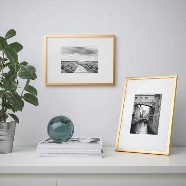 A table with a white surface displays a potted plant, a glass sphere on books, and two gold-framed black-and-white photographs, one propped upright.