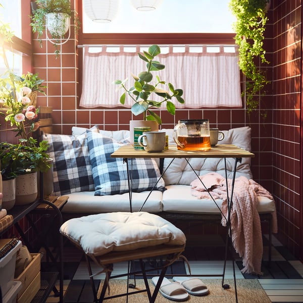 Cosy balcony with red brick wall, pink-striped MATILDA curtain, small table with tea, cushioned bench, footstool, blanket, and slippers.