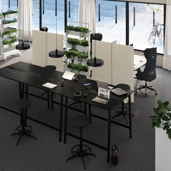 Modern coworking space withMITTZON high tables, black, featuring laptops, stools & chairs, glass partitions, hanging plants, and a large window.