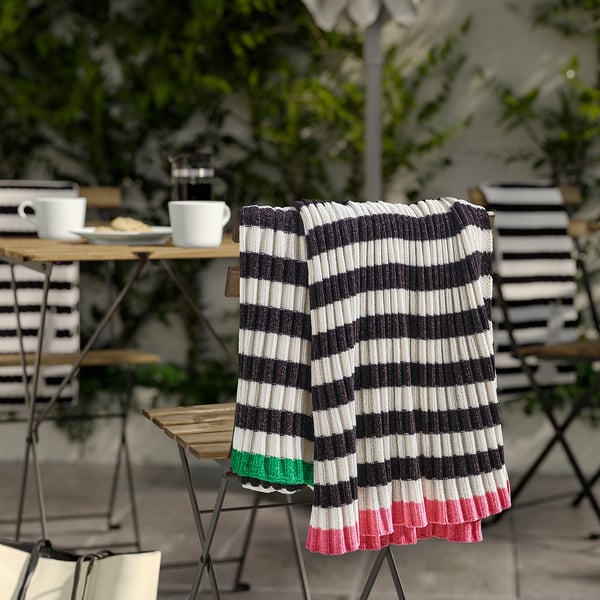 Striped MOSSMOTT throw blanket in black and white on chair, larger than mugs, amongst outdoor furniture.