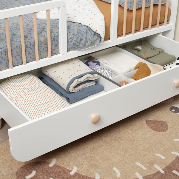 White kids cot with adjustable base and a large drawer. The drawer is open, revealing neatly folded blankets and clothes.