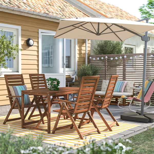 Backyard patio with NÄMMARÖ dining set, table & chairs. Beige umbrella, striped cushions. Yellow house, greenery. Cosy space.